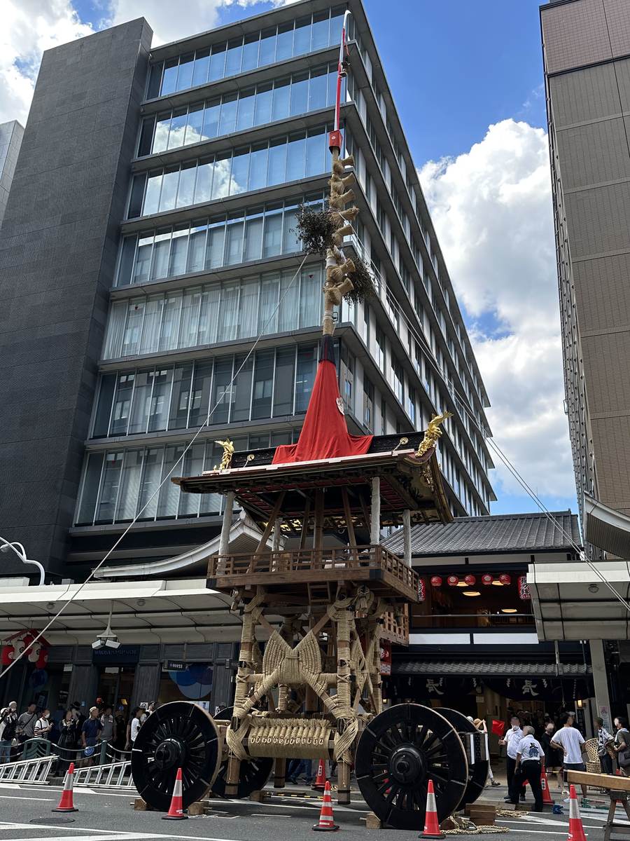 A Gion Matsuri hoko float standing in a Kyoto street with its lower body entirely bound in crossed straw rope, black wheels either side, its mast rising past the office block behind