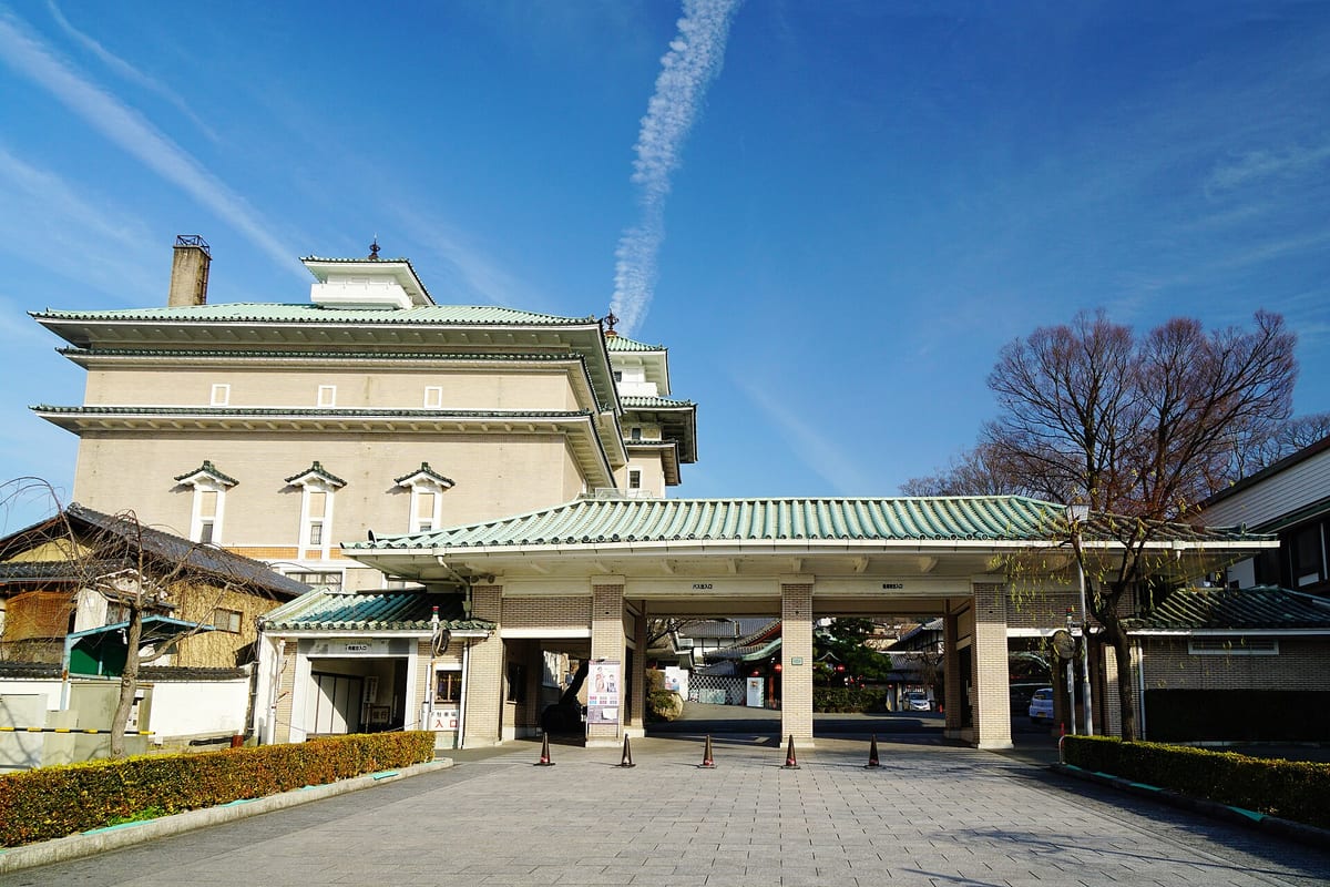 Gion Kobu Kaburenjo under a clear sky: behind, a multi-storey hall with sweeping green tiled roofs, cream walls and a small watchtower on top; in front, a matching green-tiled porte-cochère with traffic cones between its piers and an empty forecourt.