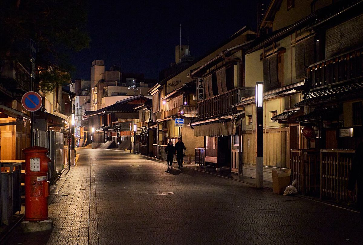 A stone-paved street in Kyoto's Gion at night, lined with two-storey wooden machiya; a long noren curtain and a white pillar lamp on the right, a red cylindrical postbox and a no-parking sign on the left, two pedestrians walking away in the middle distance
