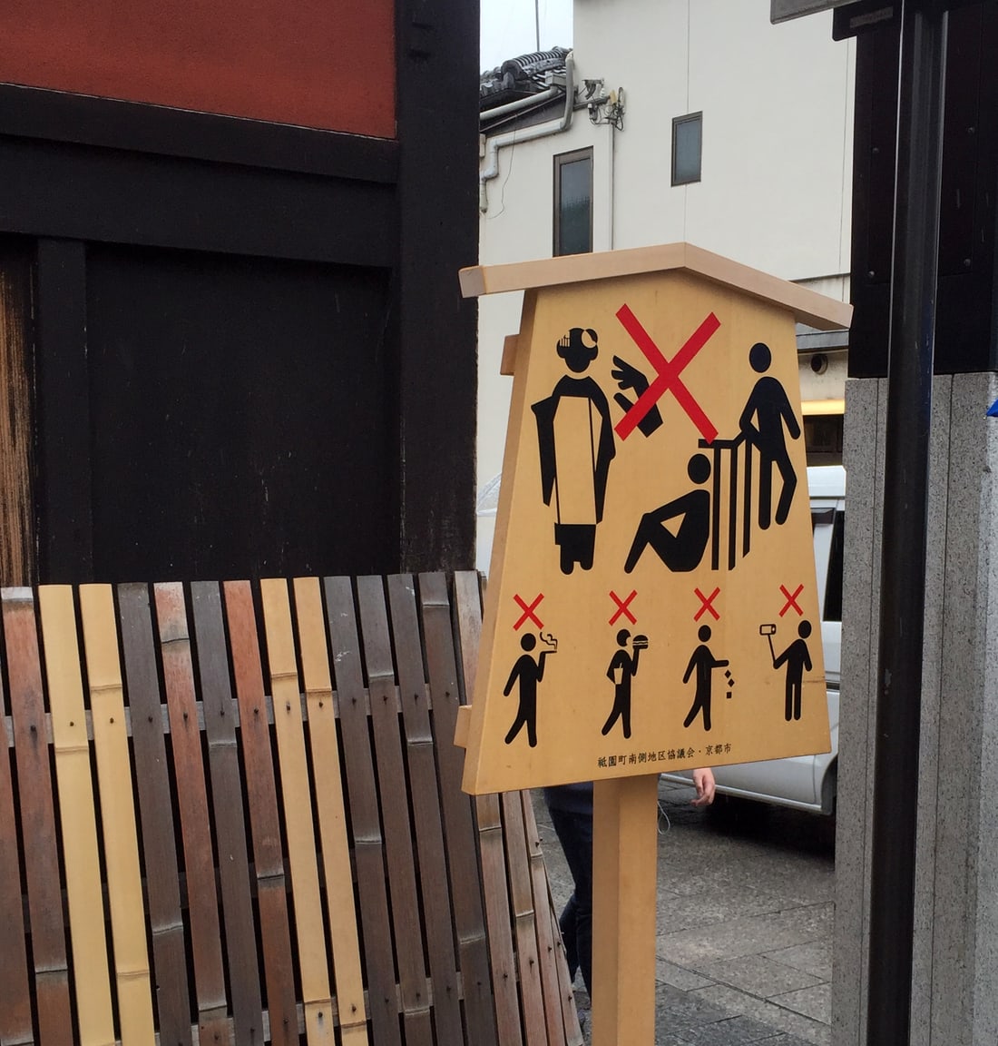 A wooden pictogram notice board on Hanamikoji Street: one large red cross strikes out both a hand reaching towards a person in kimono and a figure climbing onto a wooden lattice screen, while four smaller crosses below cover smoking, eating while walking, littering and selfie sticks; the footer credits the Gion-Machi South District Council and Kyoto City, with a komayose lattice fence to the left.