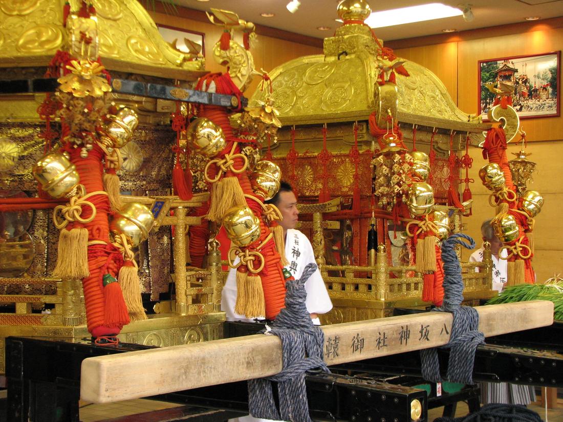 Three gilded portable shrines parked side by side indoors, their red-wrapped posts hung with brass bells, a white carrying beam in front inscribed with the name of Yasaka Shrine