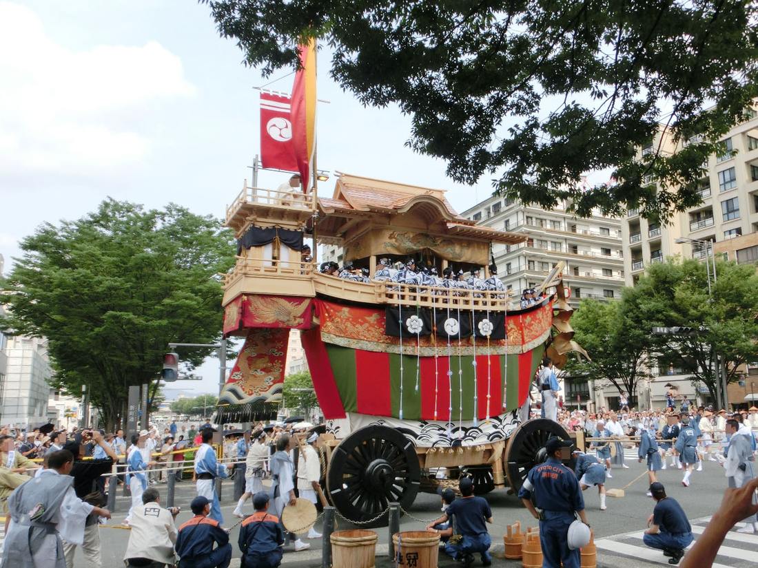 The boat-shaped Ofune-hoko standing at an Oike Street junction in Kyoto, a row of wooden buckets set out on the road in front of it and firefighters in navy uniform crouching alongside