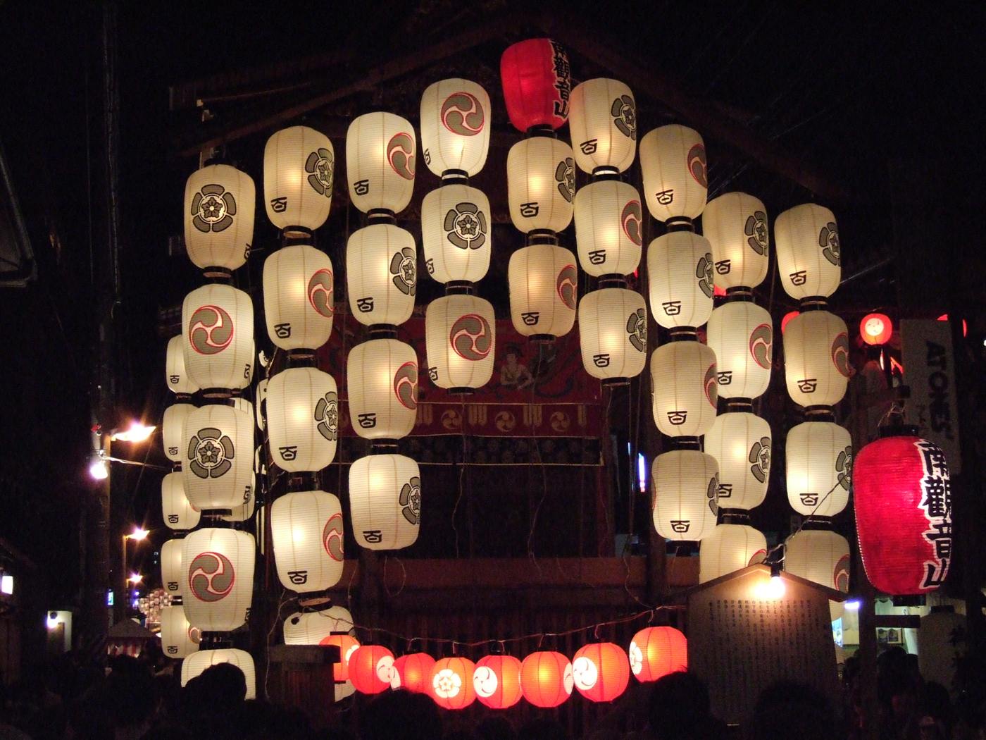 A wall of lit white paper lanterns on a Gion Matsuri float at night, a string of small red lanterns below them, and the silhouetted heads of a crowd along the bottom edge