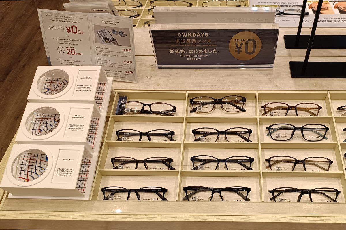 Lens option cards on an OWNDAYS frame display table in Japan, next to three progressive-lens vision samples labelled Premium, Standard and Normal