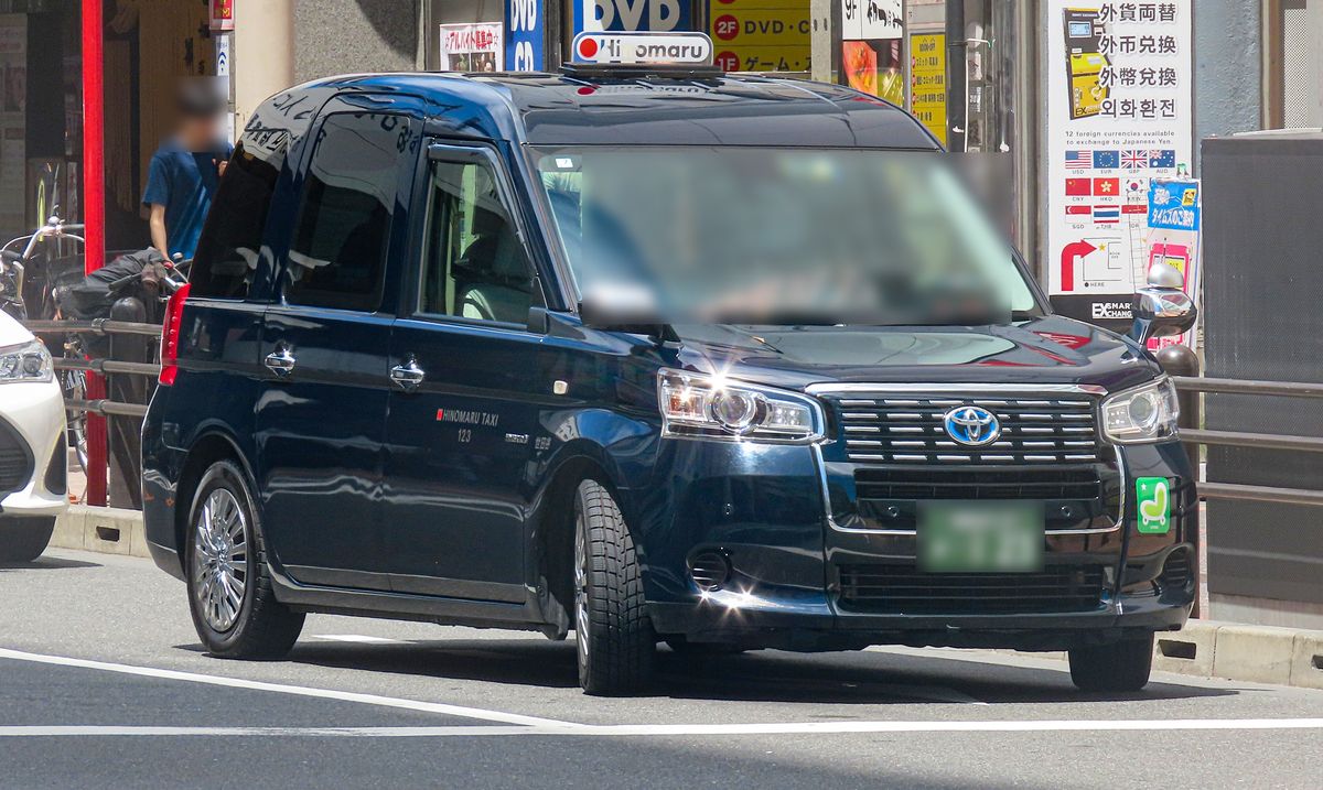 秋葉原車站南側，一台深藍色 JPN TAXI 貼著人行道護欄與路緣，車頂燈寫著 Hinomaru，背景是 DVD・CD 與遊戲店及外幣兌換的招牌