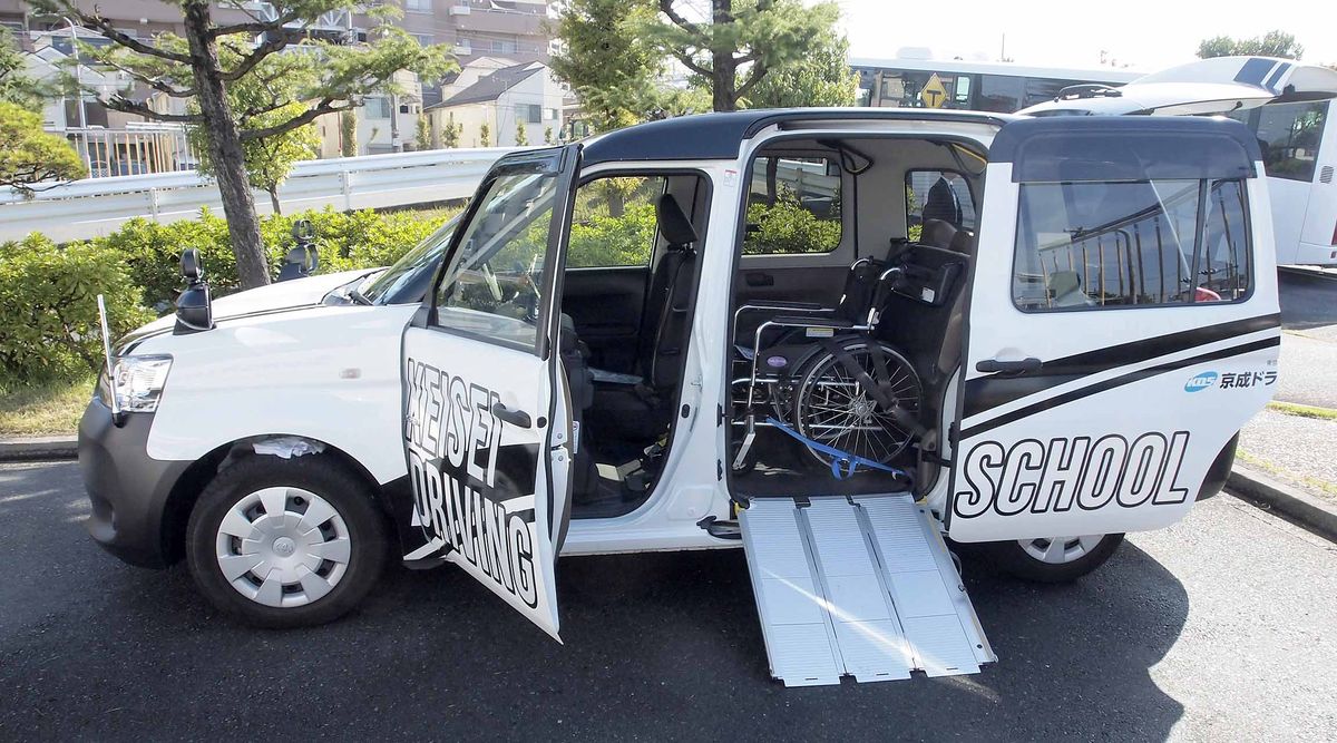 一台白色 JPN TAXI 打開前門與後側滑門，滑門下方放下鋁製斜坡板，車廂內固定著一台輪椅，車身印著「KEISEI DRIVING SCHOOL」