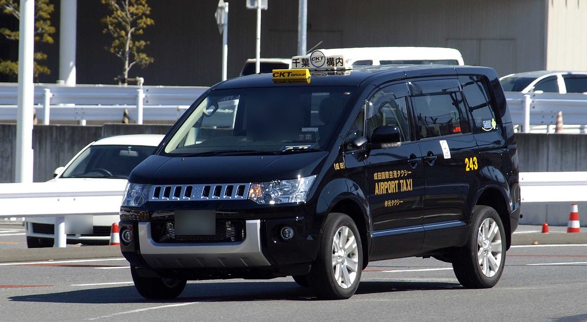 成田機場第 1 航廈前道路上的一台黑色三菱 Delica 計程車，車身印著「成田国際空港タクシー AIRPORT TAXI」與編號 243，車頂燈寫著「千葉 CKT 構内」