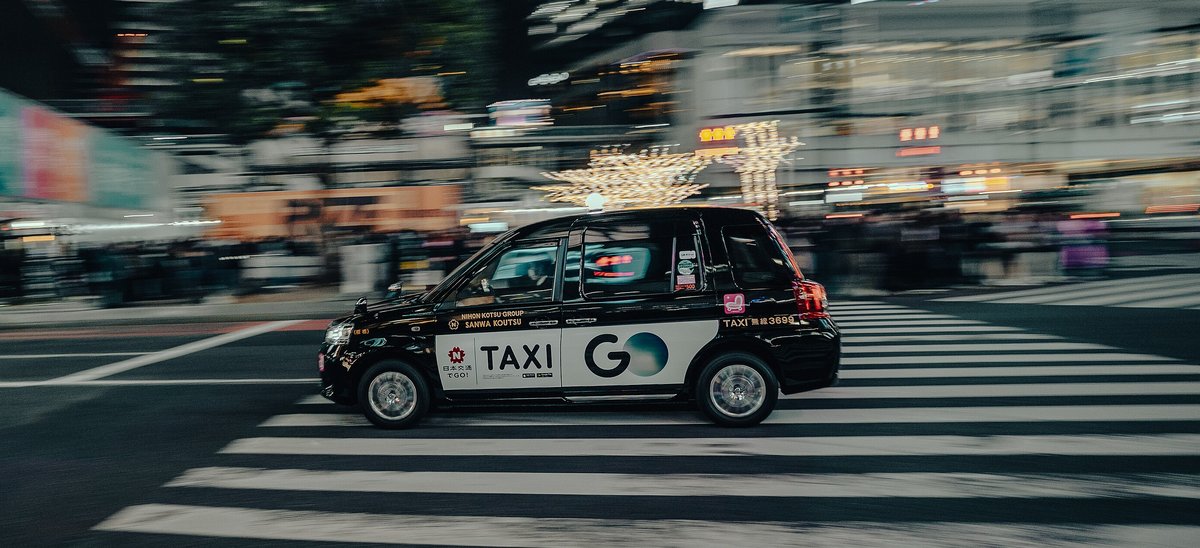 夜間的澀谷路口，一台黑色 JPN TAXI 駛過斑馬線，車門印著「TAXI GO」與日本交通集團字樣，背景的人群與霓虹燈因追焦拍攝而拉成模糊線條