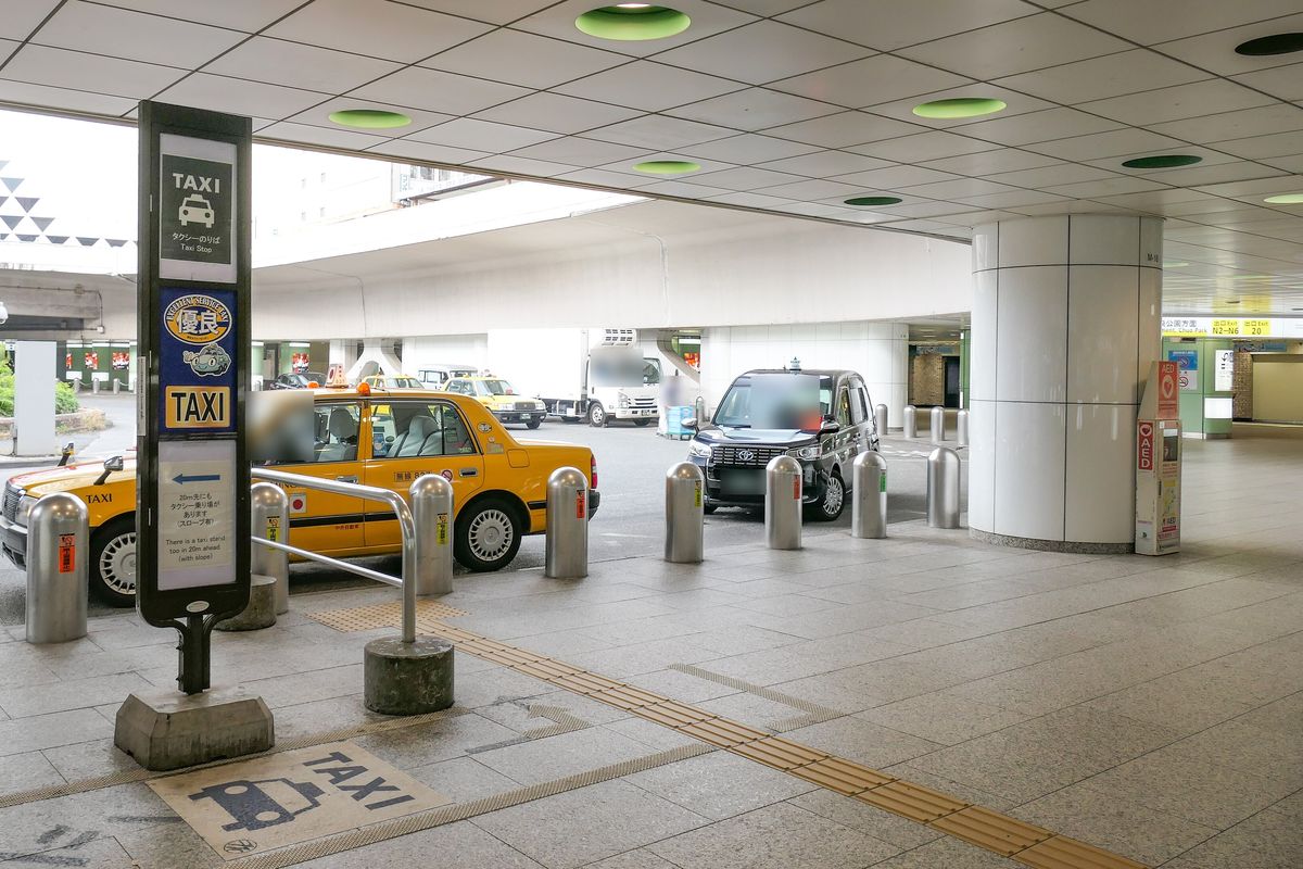 新宿車站西口的計程車招呼站，立牌寫著「TAXI タクシーのりば Taxi Stop」，地上畫著計程車圖案，路邊停著一台黃色和一台黑色計程車在等客