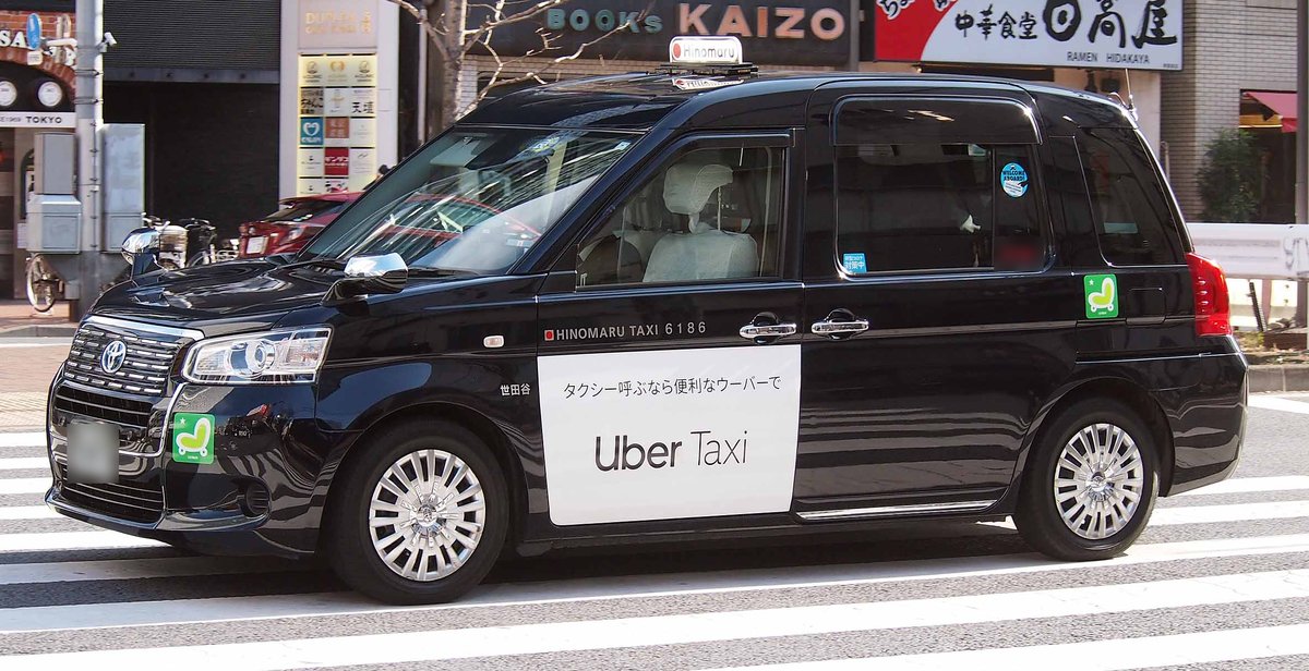 東京街頭斑馬線上一台黑色 JPN TAXI，車門貼著白底的「Uber Taxi」廣告，上面一行寫「タクシー呼ぶなら便利なウーバーで」，車身印著 HINOMARU TAXI 6186