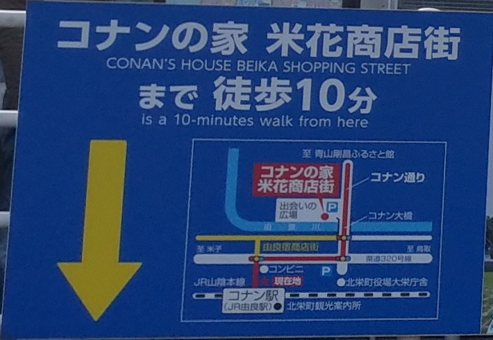 由良站前的藍色導覽牌,寫著「コナンの家 米花商店街まで徒歩10分」,下方簡圖標出コナン駅(現在地)、往北的コナン通り、跨由良川的コナン大橋、出会いの広場與米花商店街,最上方箭頭寫「至 青山剛昌ふるさと館」