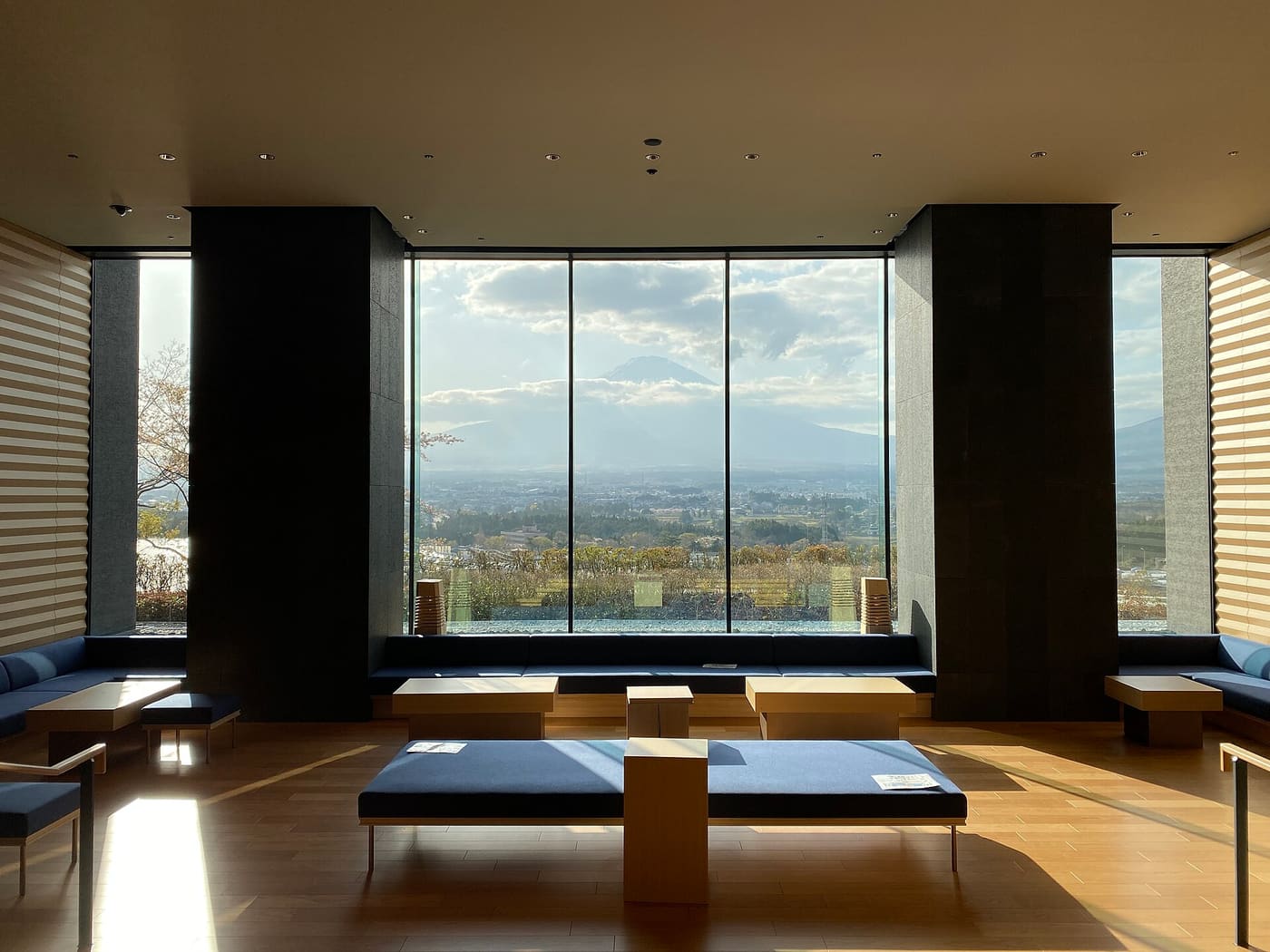 Floor-to-ceiling windows in the HOTEL CLAD lounge framing Mt. Fuji, its summit emerging above a band of cloud over the Gotemba townscape, with low blue benches and wooden tables inside