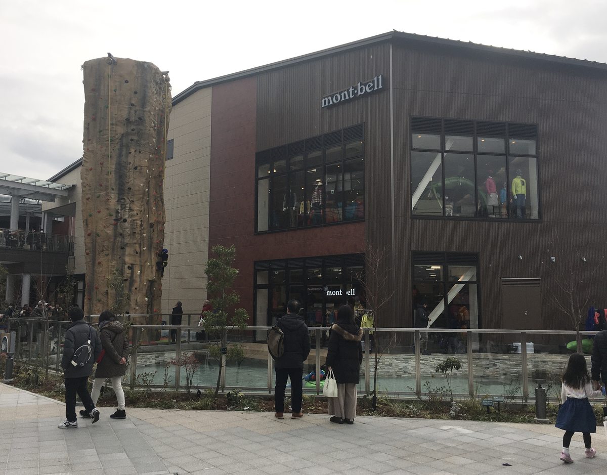 南町田グランベリーパーク的 Montbell:深褐色的兩層樓獨棟店面,外牆與一樓入口各有 mont-bell 字樣,二樓大窗裡站著穿外套的人形模特兒,左邊是一座岩壁造型的攀岩塔,店前有水池與幾位背對鏡頭的遊客