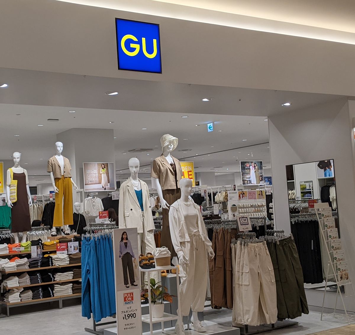 購物中心裡的 GU 門市入口：上方是藍底黃字的 GU 方形招牌，門口幾尊人形模特兒穿著米白色與卡其色的春夏服裝，旁邊是掛滿褲子的衣架與疊著上衣的層板