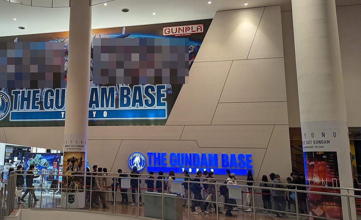 DiverCity 頂樓挑高中庭裡的 THE GUNDAM BASE TOKYO 本店入口:牆上是寫著 THE GUNDAM BASE TOKYO 與 GUNPLA 的大型看板(插圖部分被上傳者打了馬賽克),下方亮藍色的店名燈箱前,一排人沿著欄杆與紅色圍繩排隊等著進店