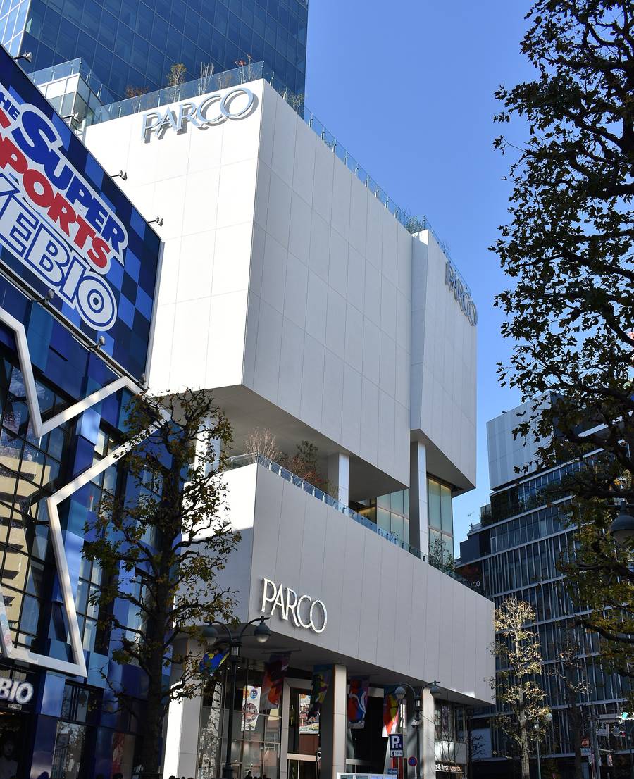 澀谷 PARCO 的外觀:白色方塊堆疊的建築,上下兩處掛著銀色 PARCO 字樣,中段有種了樹的露台,左邊是藍色外牆的運動用品店招牌,右邊是行道樹與玻璃帷幕大樓