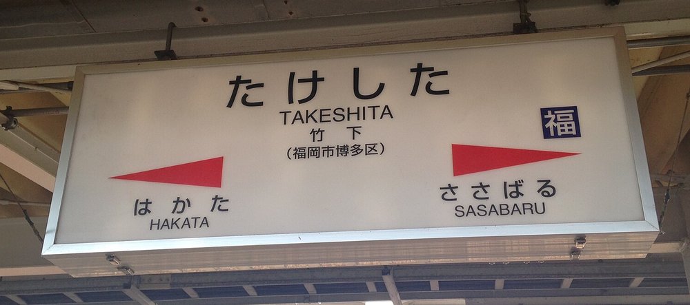 JR 竹下站月台上的白底站名牌:中間大字「たけした」與 TAKESHITA、竹下(福岡市博多区),左邊紅色箭頭指向「はかた HAKATA」,右邊紅色箭頭指向「ささばる SASABARU」,右上角有一個藍底的「福」字