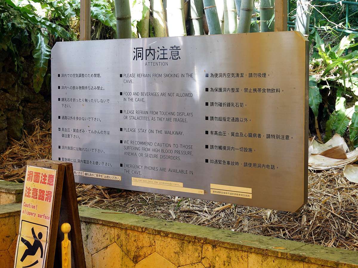 玉泉洞入口前的不鏽鋼「洞内注意」告示牌，日、英、中三語並列：洞內禁菸、禁止攜帶飲食、勿碰折鐘乳石、勿離開步道、高血壓與貧血者注意、緊急時用洞內電話；左前方立著黃底「滑面注意」的 A 字立牌