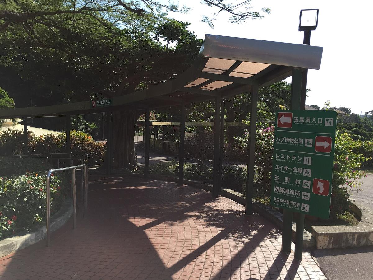 おきなわワールド園內往玉泉洞入口的有頂步道：紅磚地面、鋼架遮雨棚，綠色指標寫著左轉「玉泉洞入口」，右轉是ハブ博物公園、レストラン、エイサー会場、王国村等