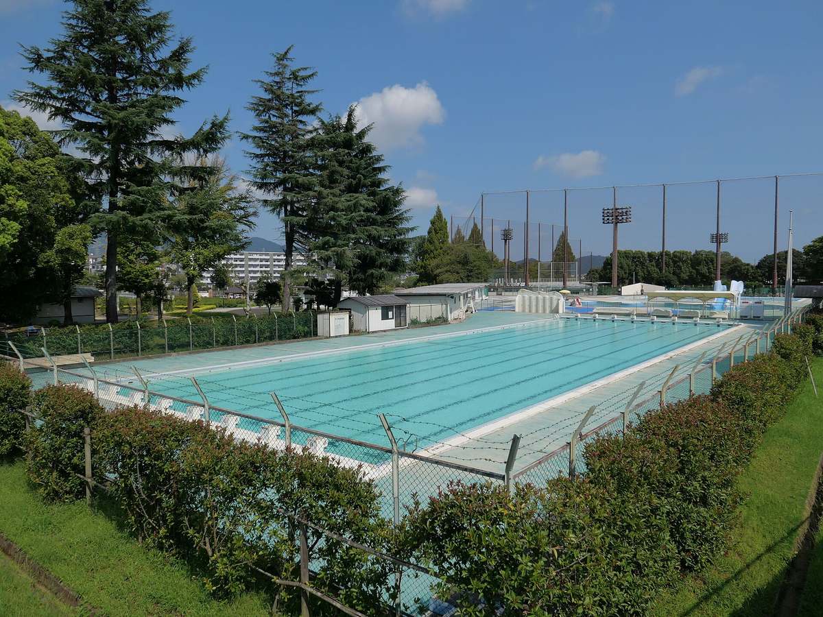 The 50-metre outdoor pool at Hadano Culture Park, Kanagawa: eight lanes of still, empty water in a rectangular basin, white tiled deck and clipped hedges around the edge, a wire fence beyond that, tall ball-park netting and floodlight masts in the distance, clear sky above