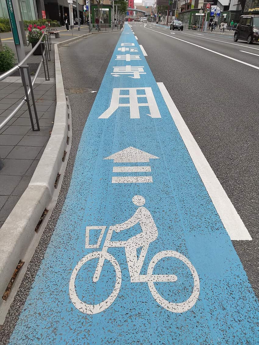 Hakata Station approach road in Fukuoka: the leftmost lane is surfaced entirely in pale blue with Japanese characters reading bicycles only painted on it, followed by a forward arrow with three chevron bars and a large bicycle pictogram, divided from the car lane by a solid white line and from the footway by concrete separator blocks, with vehicles facing the camera on the far carriageway