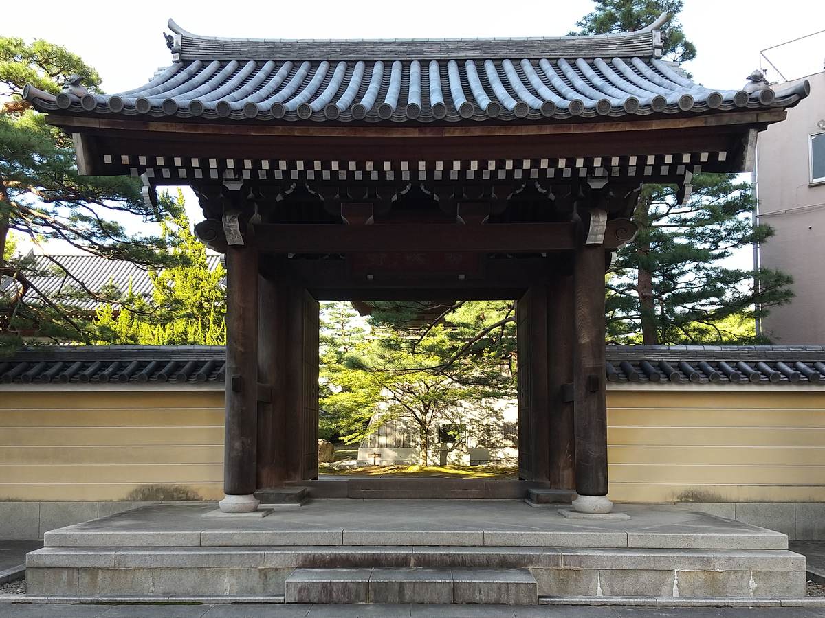 The wooden sanmon gate of Jotenji, dark timber posts under a tiled roof on a granite platform, flanked by ochre earthen walls and pines, its doors open onto trees inside