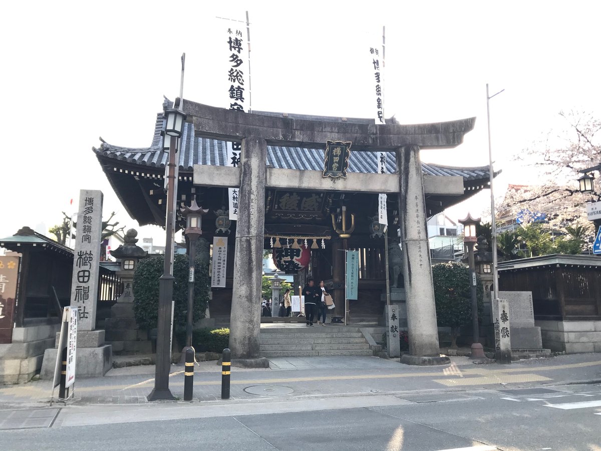 The stone torii of Kushida Shrine with the wooden two-storey gate behind it, a plaque reading Kushida-gu hung in the centre of the torii, white banners reading Hakata Sochinju on either side, and a cherry tree in blossom on the right