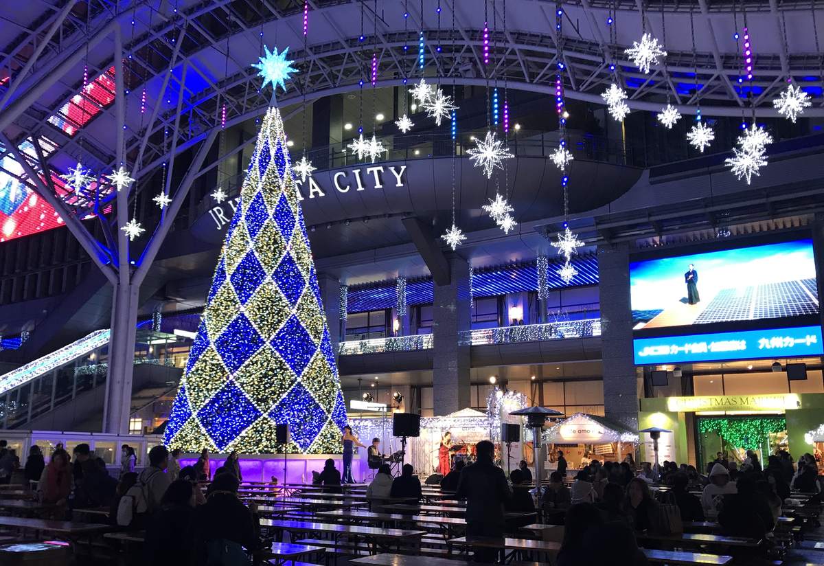 2019 年 12 月 JR 博多站前廣場會場的夜景：站房大屋簷下一座藍白菱格燈飾的大聖誕樹，後方牆上有 JR HAKATA CITY 字樣，樹腳是有人演奏的舞台，前景是成排的木製長桌與坐著的人，右側有白色屋頂的木屋攤位與寫著 CHRISTMAS MARKET 的綠色看板，頭頂垂著雪花形燈飾