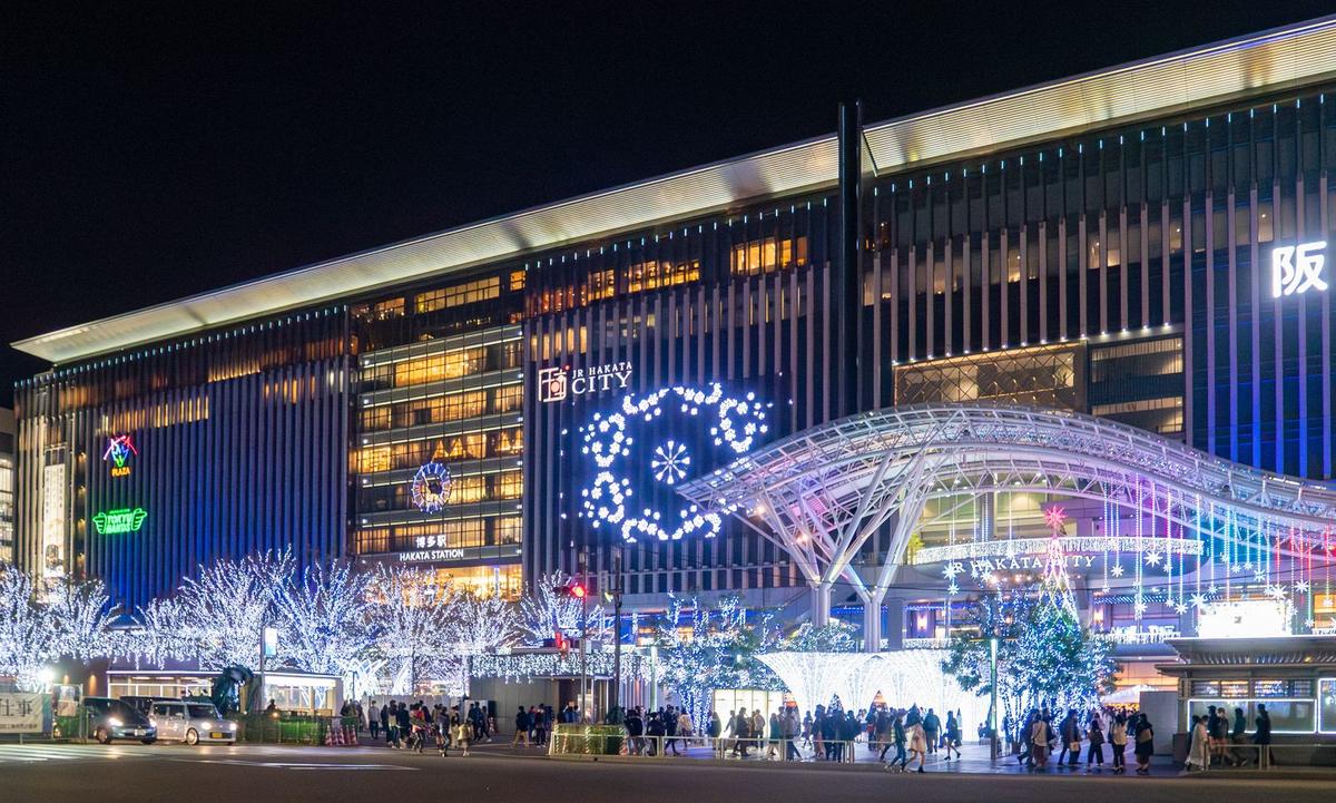 2020 年 11 月夜裡隔著馬路看 JR 博多站博多口：整面直條紋外牆的站房大樓，牆上有 JR HAKATA CITY 與「博多駅 HAKATA STATION」的字樣、一面大時鐘與藍白色的花形燈飾，站前是弧形的白色鋼構大屋簷，廣場上的行道樹纏滿藍白燈，路口有等紅燈的人群