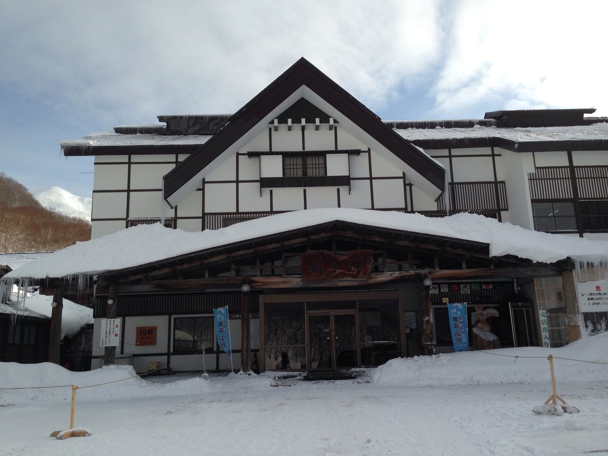 冬天的酸ヶ湯溫泉旅館正面:白牆深色木樑的三角屋頂建築,入口的木造雨棚積了厚雪、屋簷垂著冰柱,門口立著藍色旗子,地上是被踩平的雪,左後方看得到白色的山頂