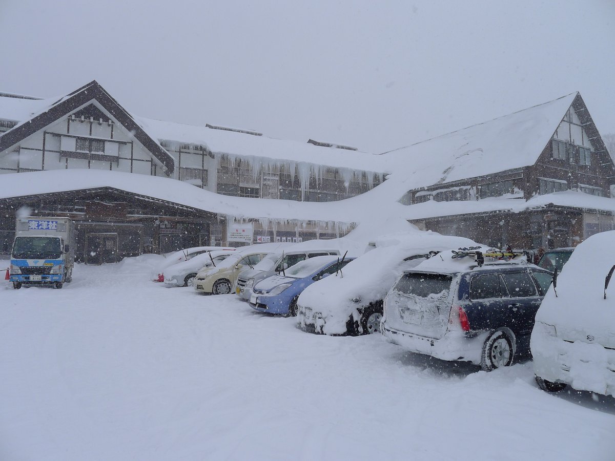12 月底大雪中的酸ヶ湯溫泉停車場:一排轎車和休旅車車頂積了厚厚一層雪,後面是屋簷掛滿冰柱的酸ヶ湯木造旅館,左邊停著一輛小貨車,空中還在下雪