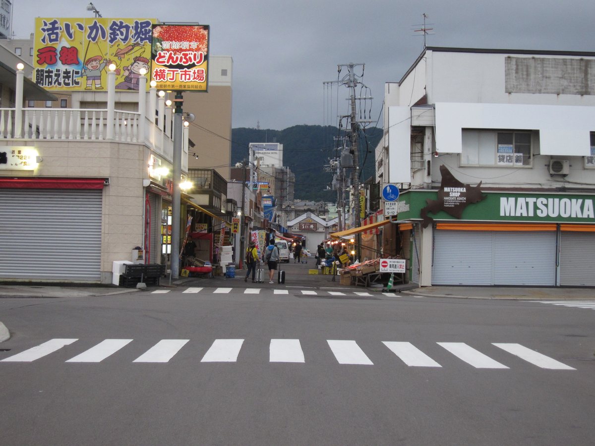 陰天清晨的函館朝市路口：前景是斑馬線，左邊大看板寫著「活いか釣堀 元祖 朝市えきに」與「函館朝市 どんぶり横丁市場」，路燈還亮著，兩側有幾間店的鐵捲門還拉著，街裡只有零星幾個人和推著的貨架