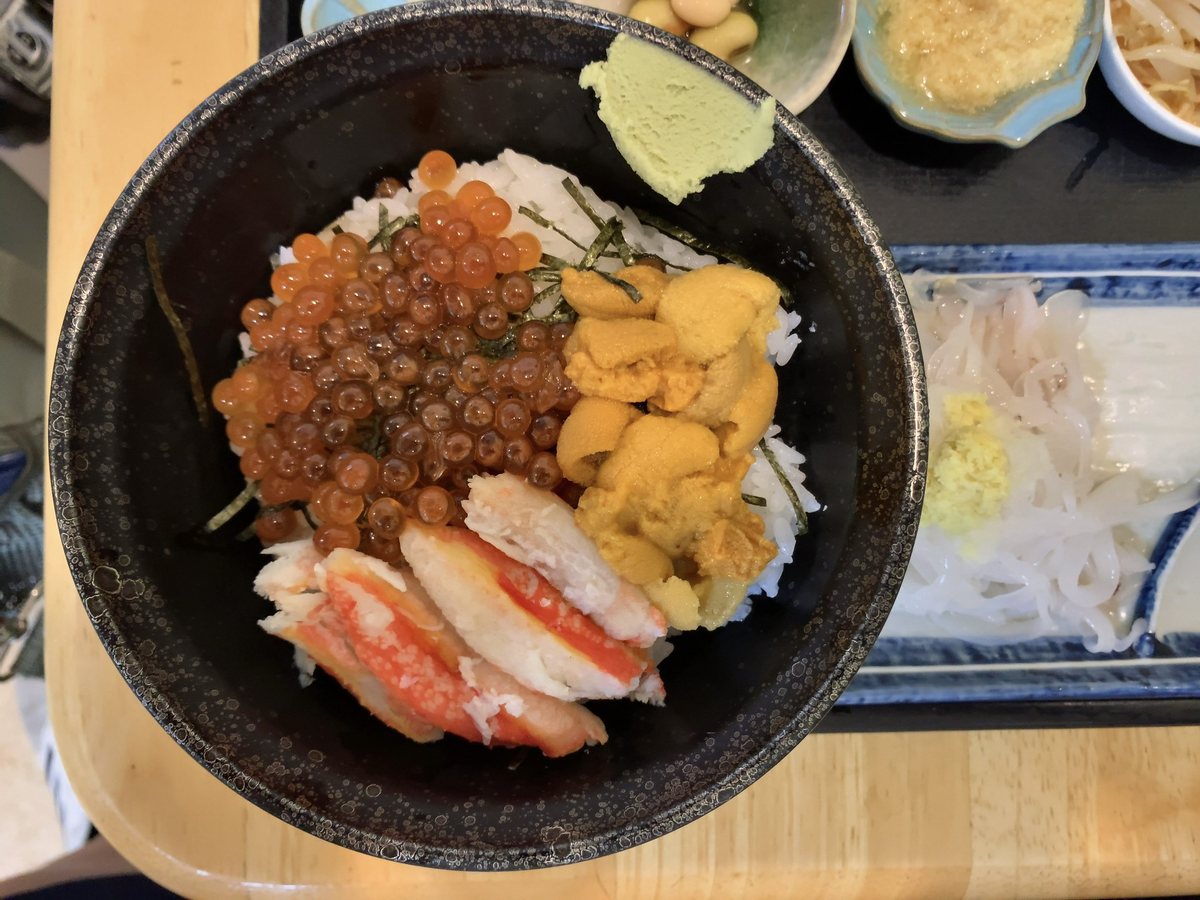 從正上方拍的一碗海鮮丼：黑色大碗裡的白飯上鋪著左半邊的鮭魚卵、右半邊的海膽，前面是幾段紅白相間的蟹腳肉，碗緣放著一小坨山葵，右邊另一個藍邊盤子上是切成細條的白色烏賊生魚片和薑泥