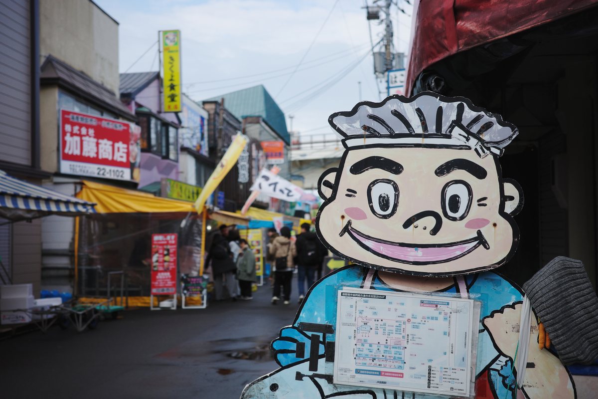 陰天的函館朝市街道，路面濕濕的有積水，左邊是「加藤商店」紅色招牌與黃色遮雨棚的攤位，遠處立著「きくよ食堂」直式招牌，幾個穿厚外套的遊客往街裡走，右前方是一個戴廚師帽的大型卡通人物立牌，胸前貼著市場的加盟店地圖