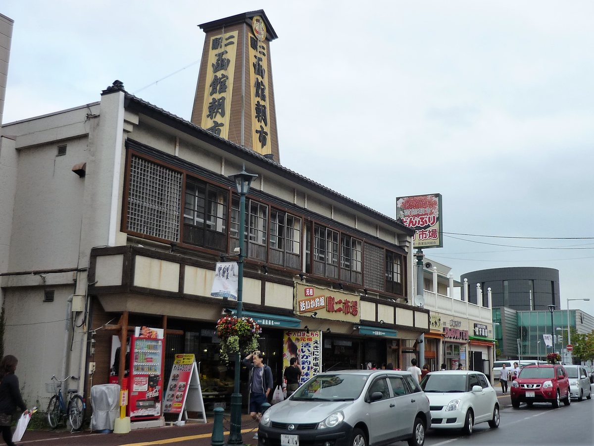 駅二市場的建築外觀：二層樓的和風木格窗建築，屋頂上立著一座直式塔狀招牌，兩面都寫著「函館朝市」，一樓招牌是「活いか釣堀 駅二市場」，右後方可以看到「どんぶり横丁市場」的招牌，前面路邊停著幾輛車