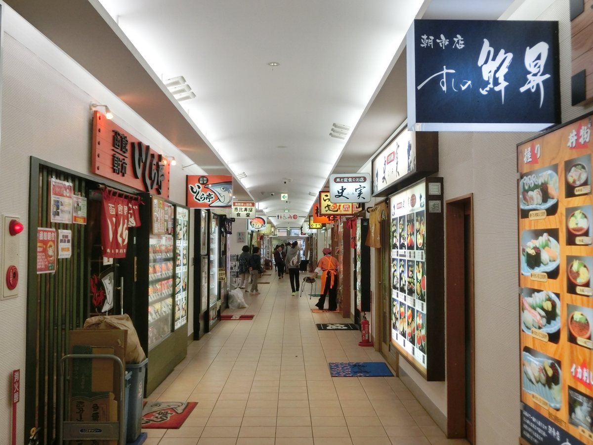 どんぶり横丁市場另一段室內走道：右邊掛著深藍底白字的「すしの鮮昇」招牌和一整片握壽司、丼飯照片的菜單板，走道中間上方是「史実」的白色招牌，左邊是「いくら亭」的紅色招牌與暖簾，遠處一位穿橘色圍裙的店員站在門口