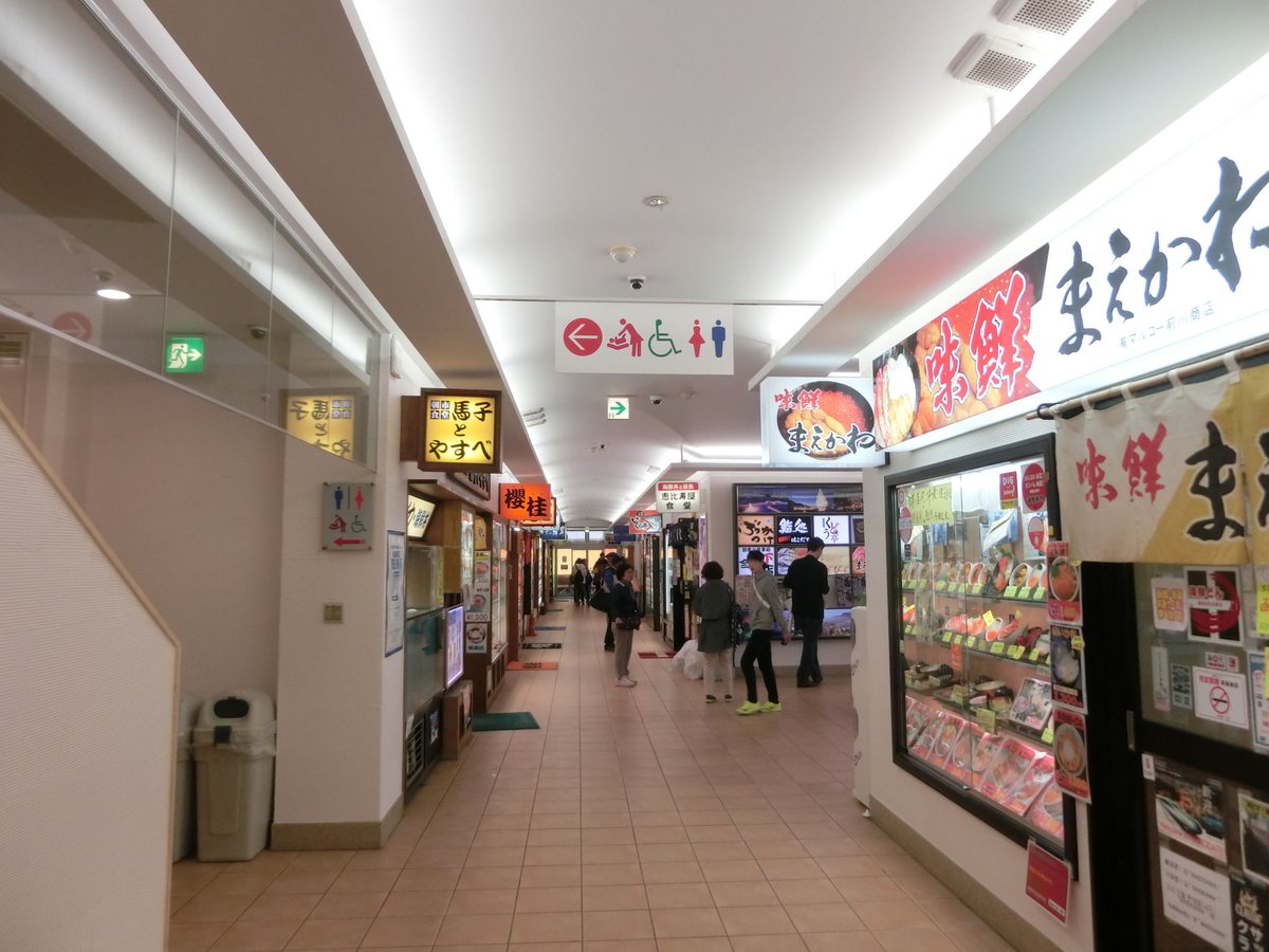 どんぶり横丁市場的室內走道：米色地磚的長廊兩側是一間接一間的食堂，右邊是「味鮮 まえかわ」的大招牌和擺滿海鮮丼樣品的櫥窗，左邊依序是「馬子とやすべ」「櫻桂」的燈箱招牌，天花板掛著指向廁所與無障礙設施的標示，走道裡有幾個遊客在看店