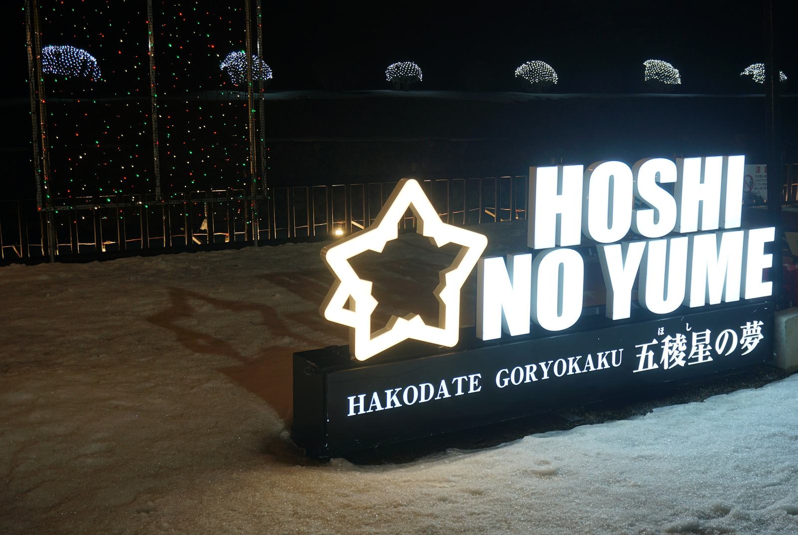雪地上一座發白光的立體字看板，左邊是星形框，右邊是大字「HOSHI NO YUME」，下方黑底寫著「HAKODATE GORYOKAKU 五稜星の夢」；看板後方是柵欄與護城河對岸的土堤，堤上一排灌木被網狀白燈包成圓球，左側有一座掛滿紅綠小燈的鐵架