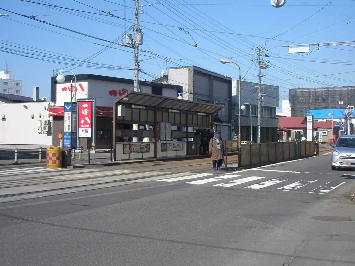 晴天的湯の川温泉電停：馬路中央一座有屋頂的候車亭，側面貼著兩塊溫泉飯店的廣告，幾個人在亭下等車，臉已被打上馬賽克；左邊立著直式站名柱，寫著「湯の川温泉」，旁邊是居酒屋「つぼ八」的紅色直式招牌，前景是斑馬線與軌道
