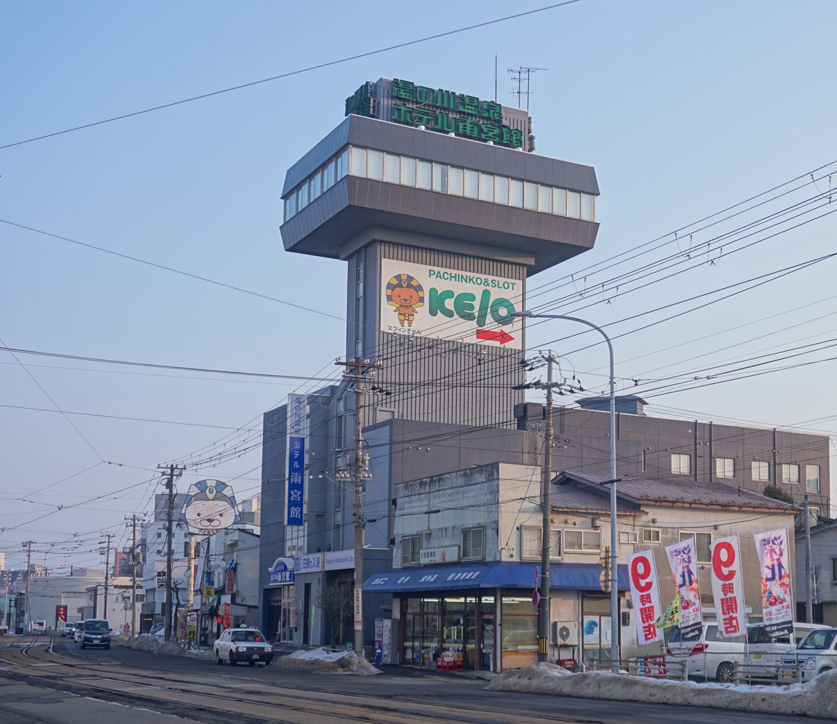 冬天傍晚的湯之川電車路：前景是市電的軌道與路邊殘雪，中間一棟大樓頂上是突出的展望樓層，屋頂綠色招牌寫「湯の川温泉 ホテル雨宮館」，樓身掛著柏青哥店的看板，右邊是一排插著廣告旗的低矮店面，左邊路上有一台計程車與幾台車，天色是淡藍帶粉紅