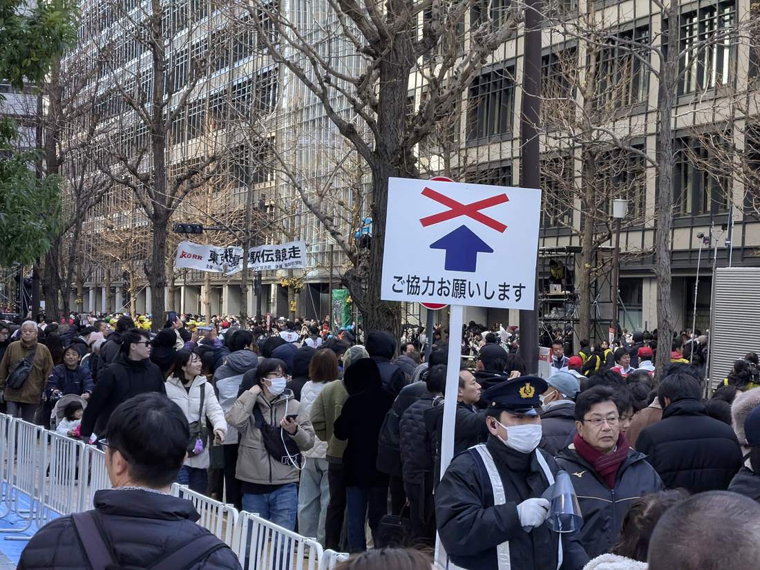 A street lined with office towers packed with spectators held behind white steel barricades on the pavement; a white sign in the centre shows a blue arrow struck through with a red cross above Japanese text asking for cooperation, a police officer in a peaked cap and mask stands beneath it holding a megaphone, a banner reading Tokyo-Hakone Round-Trip University Ekiden Race hangs to the left, and the street trees are bare