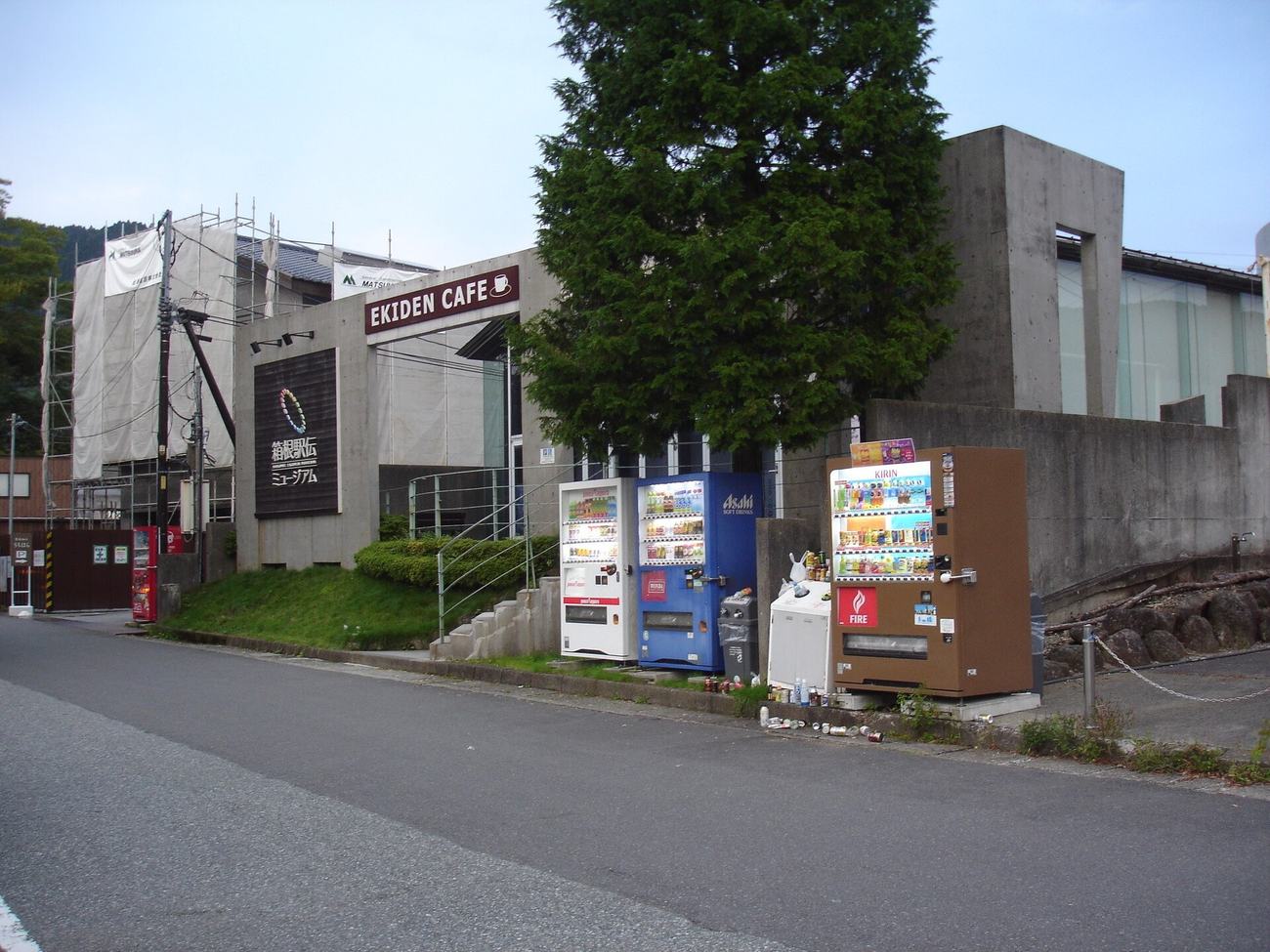 A board-marked concrete building beside a road, with a dark sign on its wall carrying a multicoloured ring logo and the Japanese name of the Hakone Ekiden Museum, an EKIDEN CAFE sign above the entrance, scaffolding and white sheeting covering the left half of the building, four drink vending machines lined up at the kerb, and an empty stretch of asphalt in the foreground
