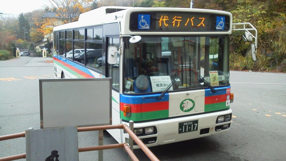 早雲山站前停著一輛白色路線巴士,車身是藍紅綠三色條紋與獅子標誌,車頭跑馬燈打著橘色的「代行バス」,擋風玻璃後立著「箱根ロープウェイ 代行バス 桃源台行」的牌子,背景是秋天轉黃的樹(2015 年 11 月拍攝)