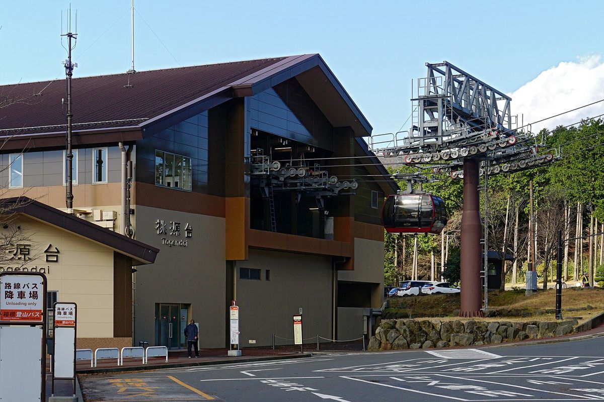 桃源台站外觀:咖啡色斜屋頂的站房,牆上寫著「桃源台 Togendai」,站房二樓伸出纜車的鋼架與滑輪,一台紅色吊廂正從右側出站;左下角是兩塊「路線バス 降車場 箱根登山バス」的站牌(2016 年 12 月拍攝)
