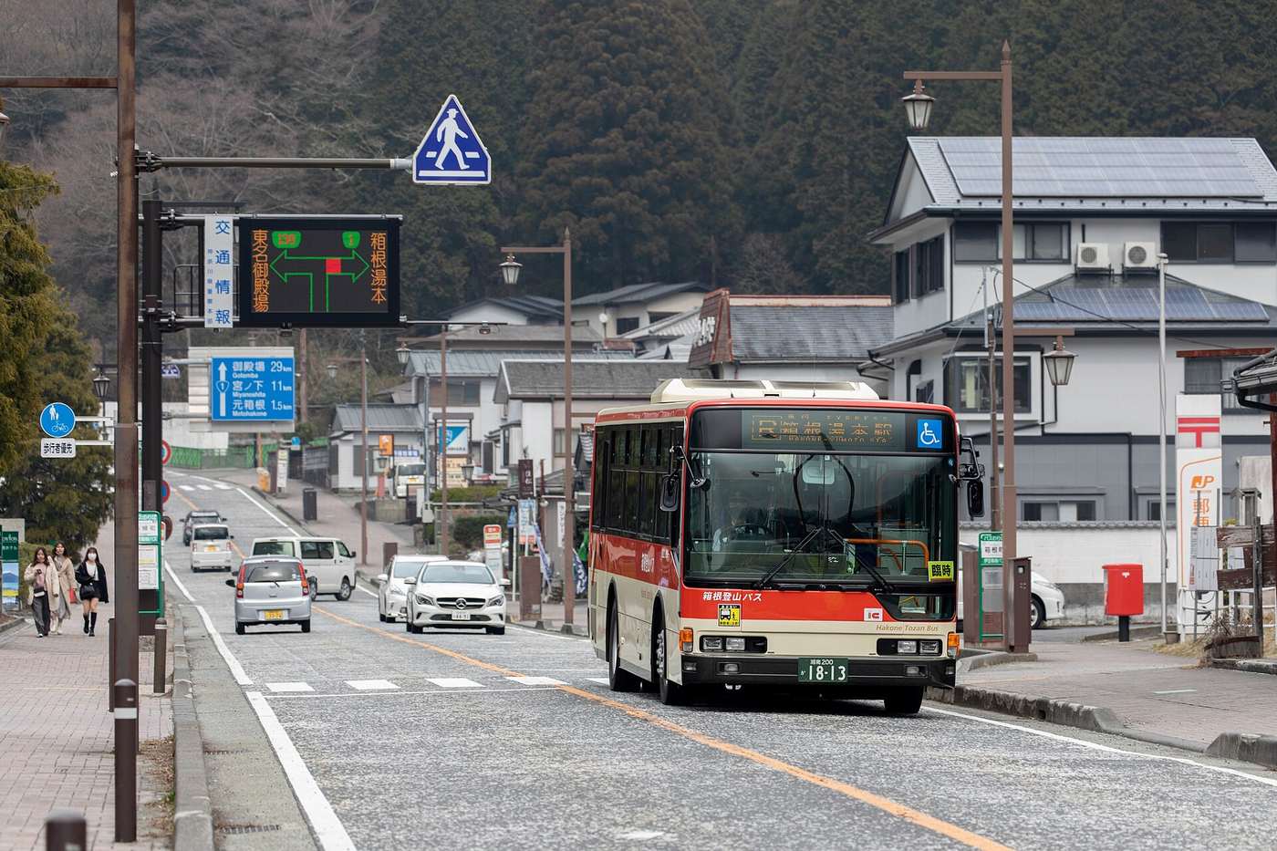 A red and cream Hakone Tozan Bus approaching head-on, its destination blind reading Hakone-Yumoto Station with an orange extra-service placard in the lower right of the windscreen; a black electronic board headed Traffic Information spans the road above it, showing green schematic routes towards Tomei Gotemba and Hakone-Yumoto with one segment lit red at the junction; a blue direction sign on the left reads Gotemba 29 km, Miyanoshita 1.1 km, Motohakone 1.5 km, and a post office mark with a red postbox stands in front of a house on the right