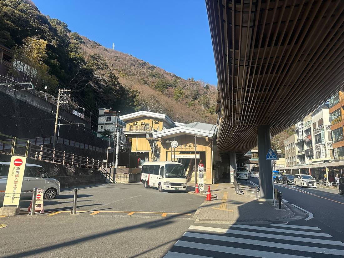 The forecourt of Hakone-Yumoto Station on a clear winter day: a cream station building with a round clock and a sign reading Hakone-Yumoto Sta., a white minibus and several orange traffic cones in the turning circle, a yellow standing board reading No Entry for vehicles at the left, a wooden-louvred pedestrian deck canopy at the upper right, and bare brown hillside with shopfronts behind