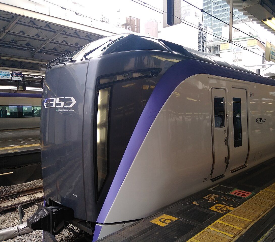 新宿站月台上停著一列特急あずさ的 E353 系列車:車頭是深灰色、側面白色車身,一道紫色斜線從車頭往後劃開,車頭與車身印著 E353 字樣,車門旁的月台地面有黃色導盲磚與乘車位置標示,頭上是月台雨棚與幾塊吊掛的指示牌,背景是新宿的高樓