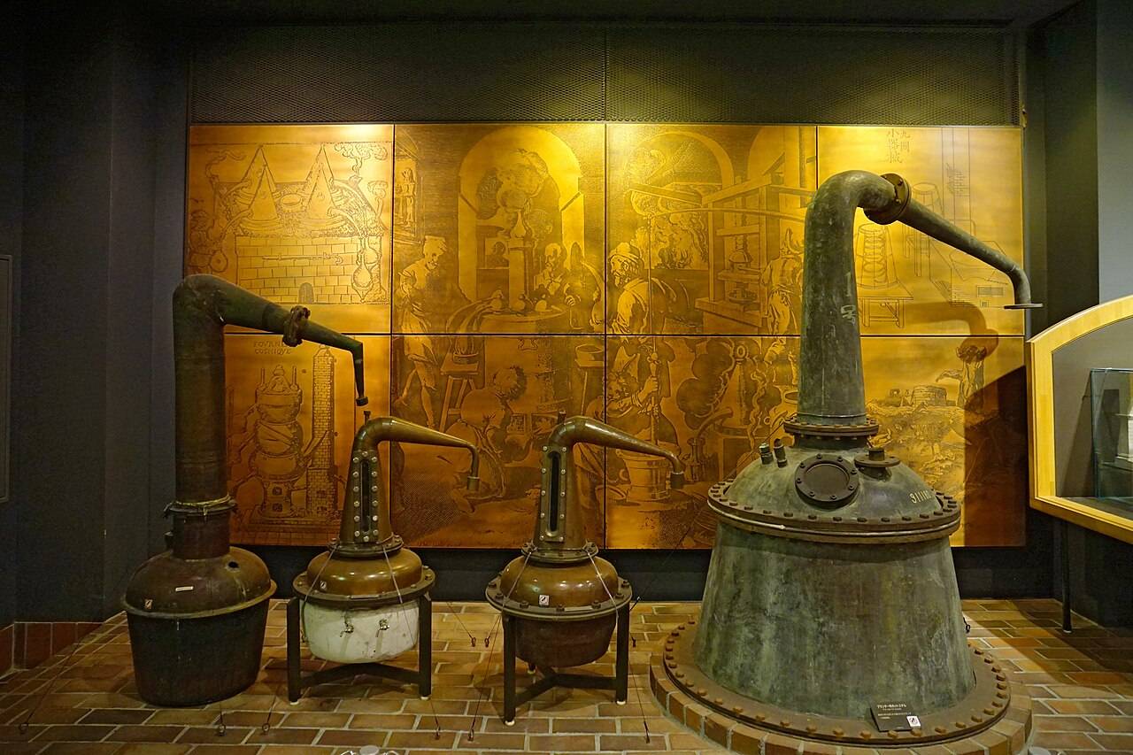 威士忌博物館館內的展示:紅磚地台上並排四座舊式蒸餾器,左邊一座高瘦、中間兩座小型的放在鐵腳架上,右邊一座是表面泛綠的大型銅釜,鍋頸彎出一根長管;背後整面金色的銅版畫牆,刻著古代蒸餾作坊的場景,畫面裡沒有人