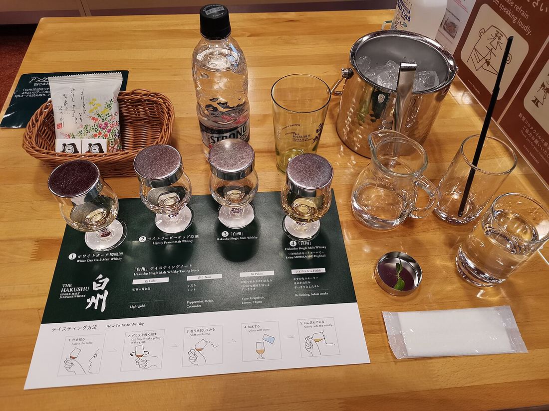 木桌上的白州試飲組:深綠色的試飲墊上排著四杯加了金屬蓋的鬱金香杯,墊上印著 ①ホワイトオーク樽原酒、②ライトリーピーテッド原酒、③「白州」、④「白州」森香るハイボール,下方是五個步驟的品飲方法圖;右邊有一桶冰塊、一壺水、兩個空杯和一小碟薄荷葉,左邊一籃小點心與一瓶氣泡水,畫面裡沒有人
