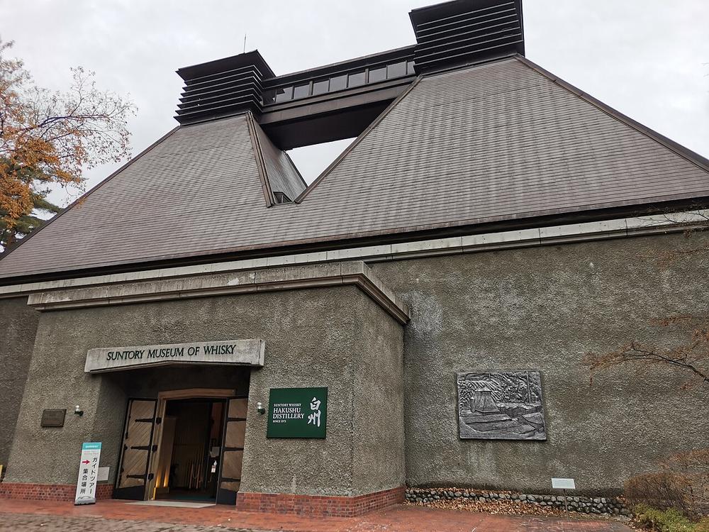 白州的威士忌博物館外觀:灰色粗糙外牆的建築,屋頂是兩座像麥芽窯的尖錐形斜頂,頂端各有一座百葉窗式的通風塔,中間用一段玻璃連廊接起來;正門上方的橫匾寫 SUNTORY MUSEUM OF WHISKY,門邊掛著綠色「白州 HAKUSHU DISTILLERY SINCE 1973」招牌,右邊牆上嵌著一塊浮雕,左邊一棵轉黃的樹,陰天,畫面裡沒有人